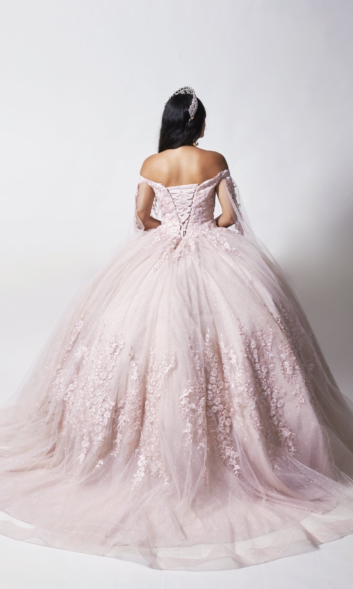 A woman in the Nina Canacci Q109 long quinceanera dress, featuring an off-the-shoulder pale pink ball gown with a floral bodice and tiara, stands facing away against a plain white background.